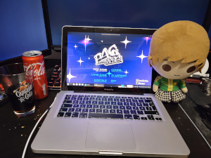 A 2012 Macbook Pro running Persona 4 Golden on Linux Mint, along with a class of cola and a Chie plushie.