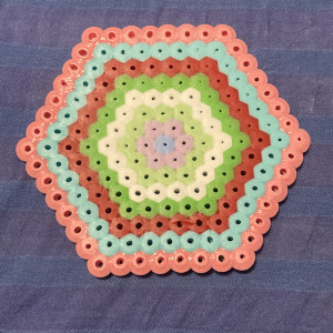 A fuse bead hexagon, with seven layers, and then an innermost bead, with the following colours, from the outside in: pink, light blue, red, green, white, light green, purple, blue.